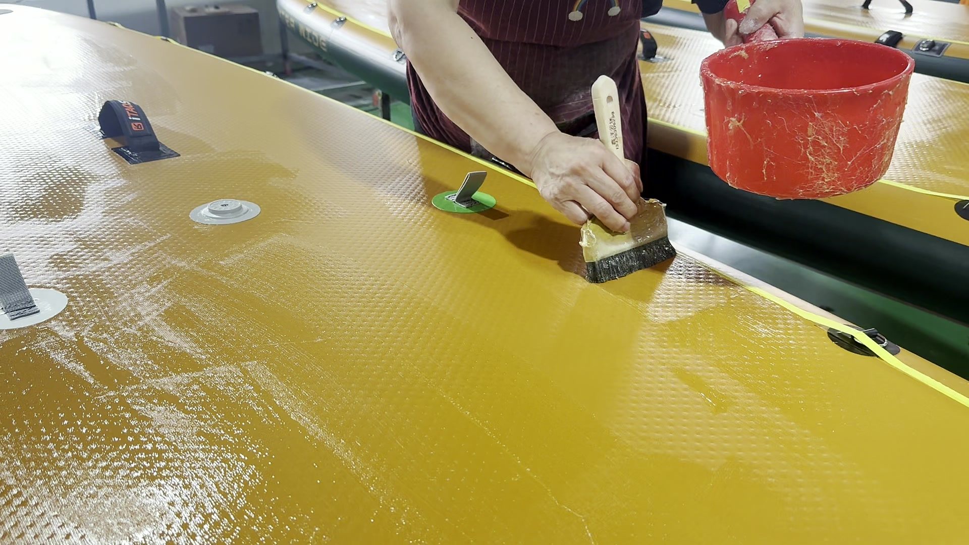 Worker brushing adhesive onto a yellow paddle board deck.